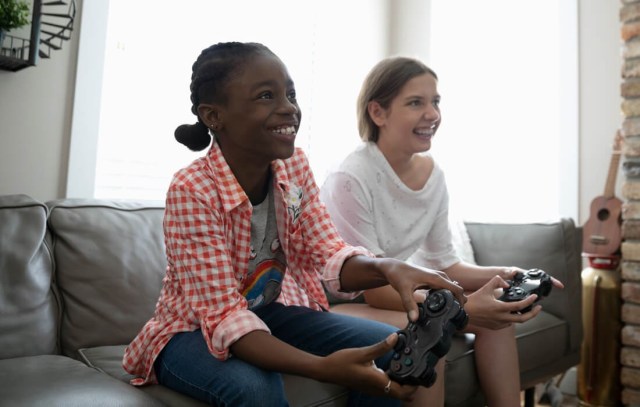 Two girls sit on the couch playing video games with controllers in each hand